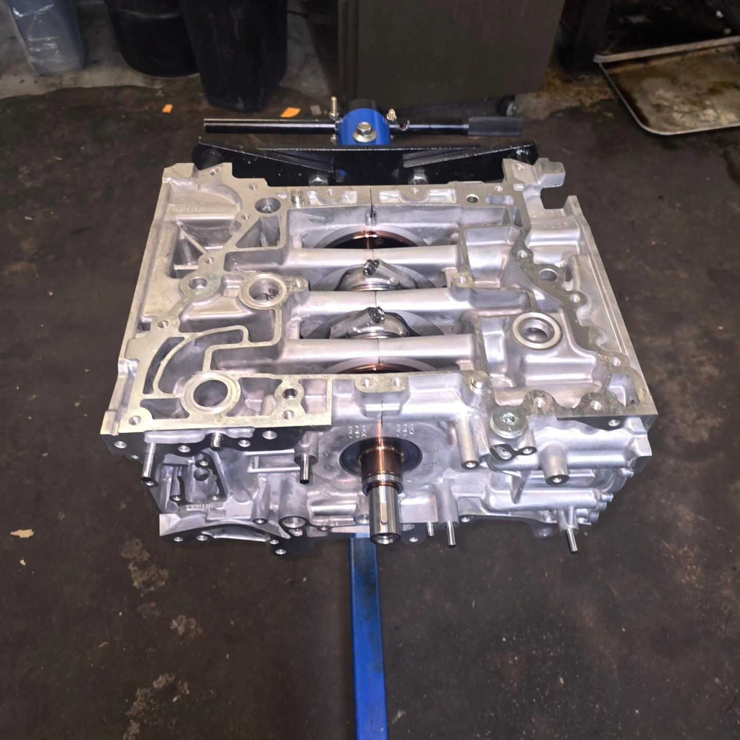 Engine block on a stand, viewed from above, in a garage. Silver metal with cylinders visible.