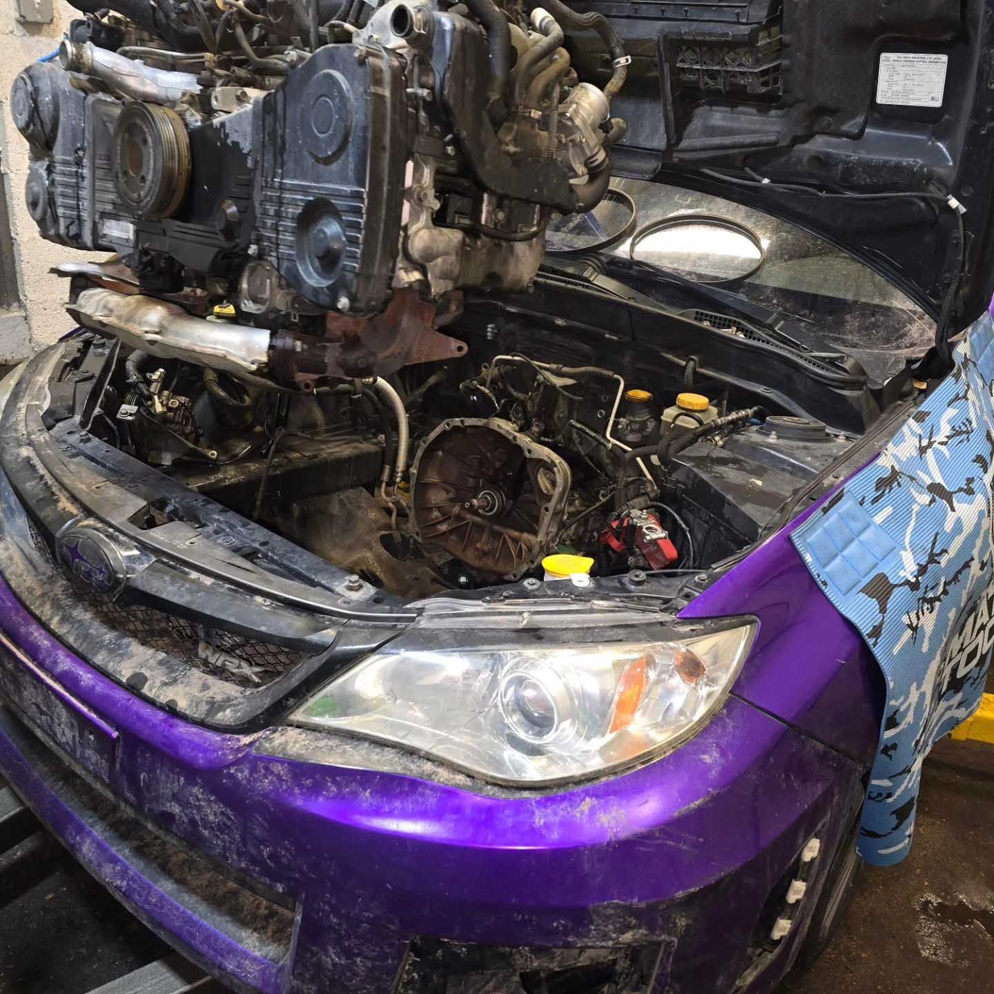 Purple car with engine removed, hood open in a repair shop.