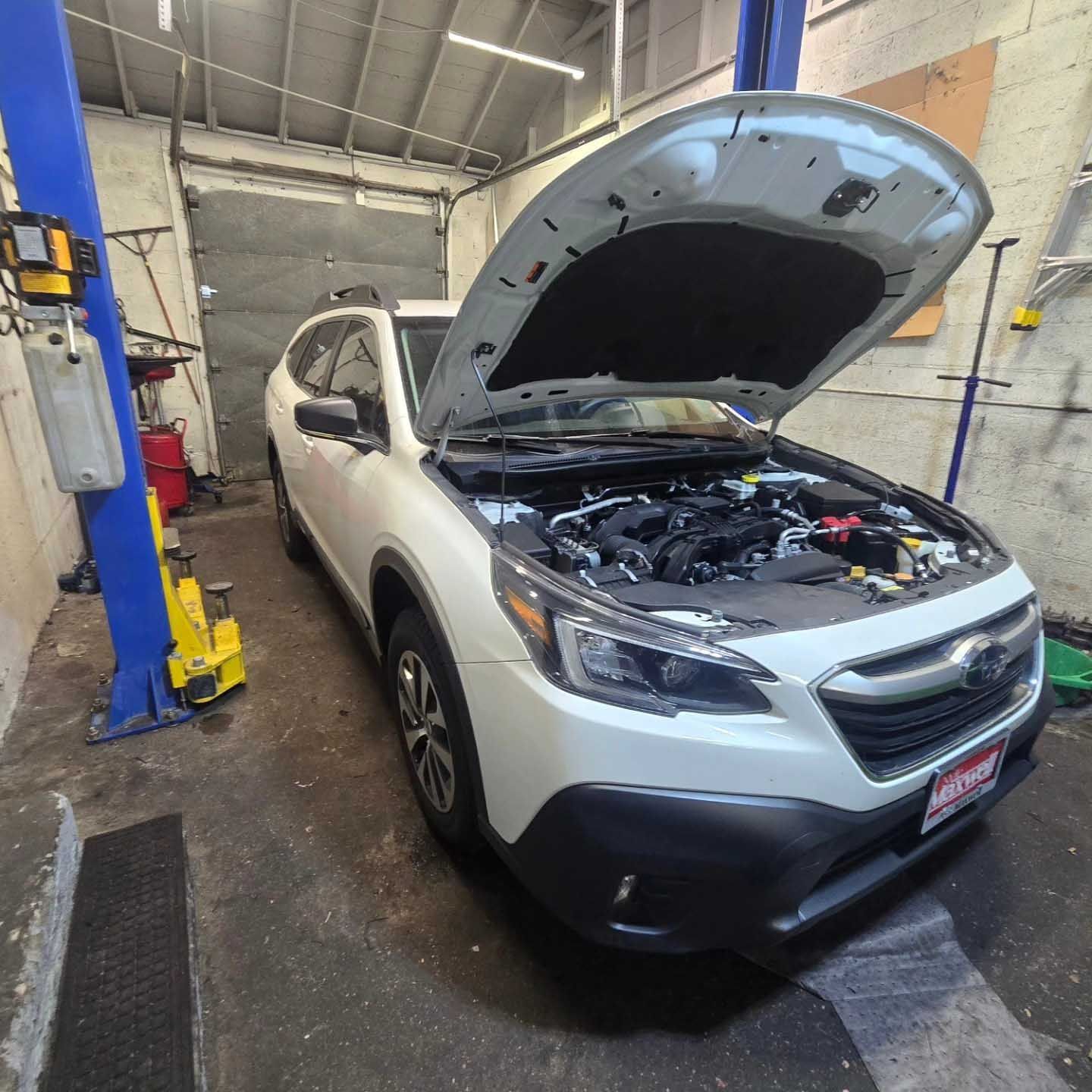 White Subaru Outback with hood open in a garage, next to a blue lift.