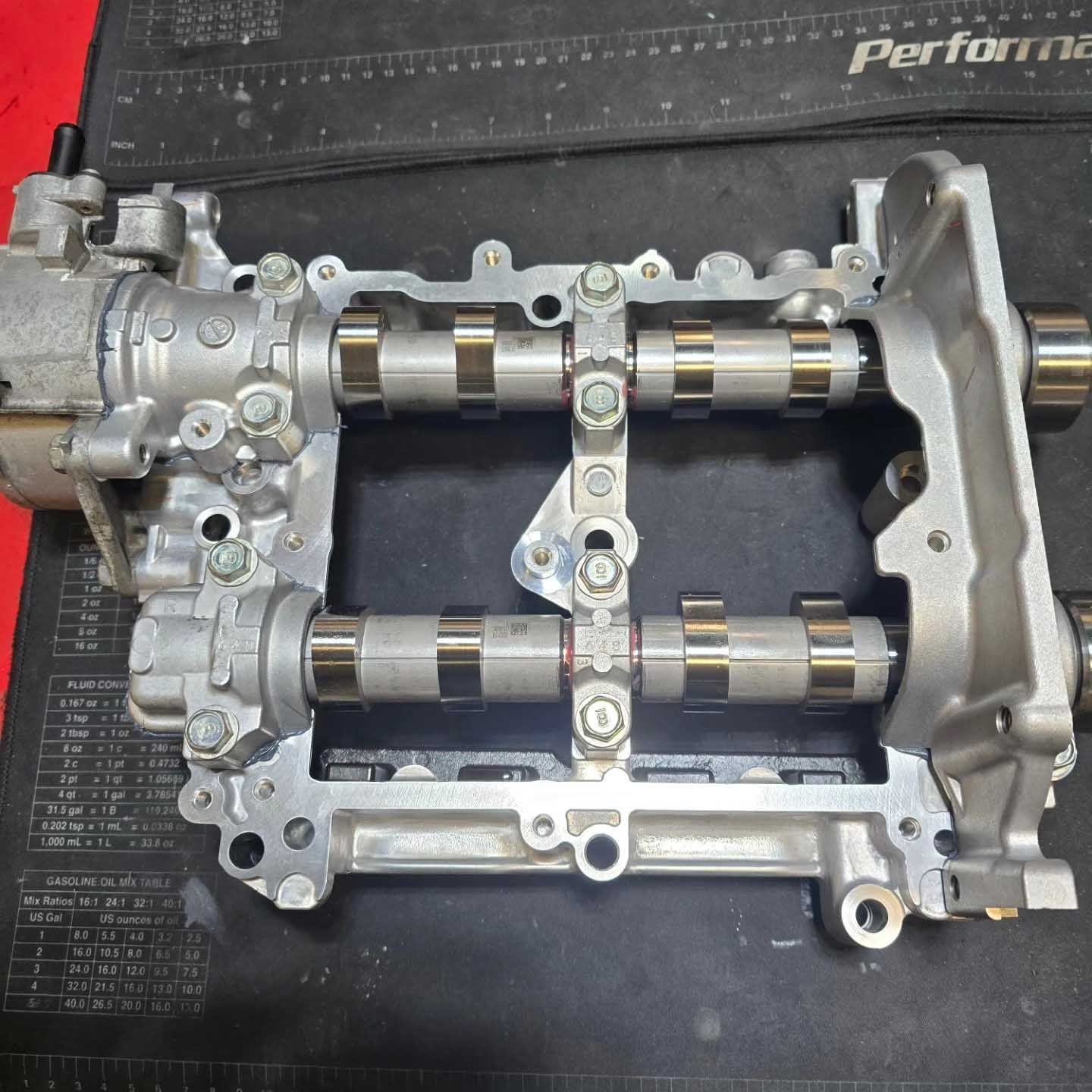 Engine cylinder head with camshafts, valves, and oil pump. Gray metal on a black surface.