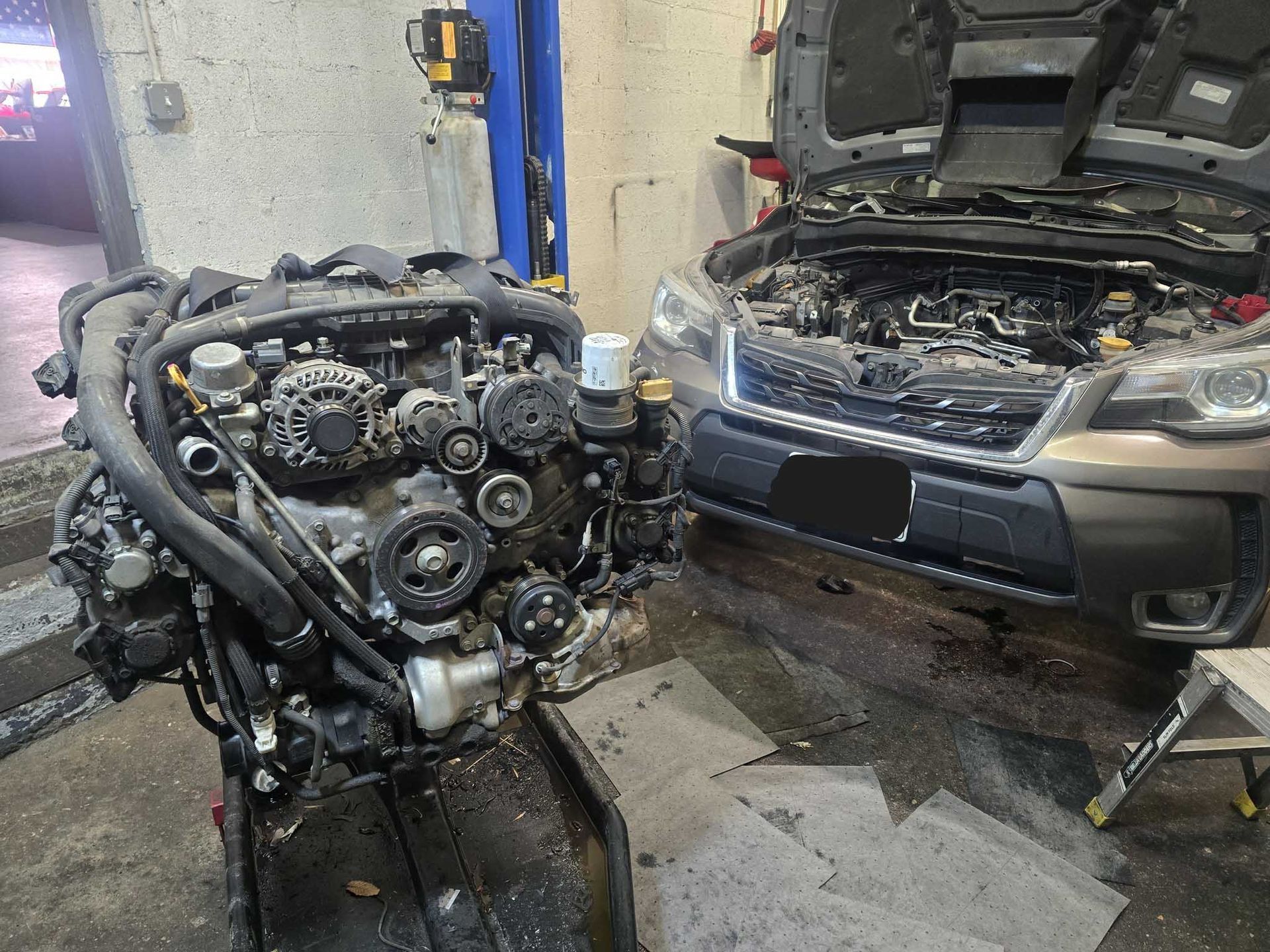 Car engine next to a Subaru with hood open in a garage, suggesting engine repair.
