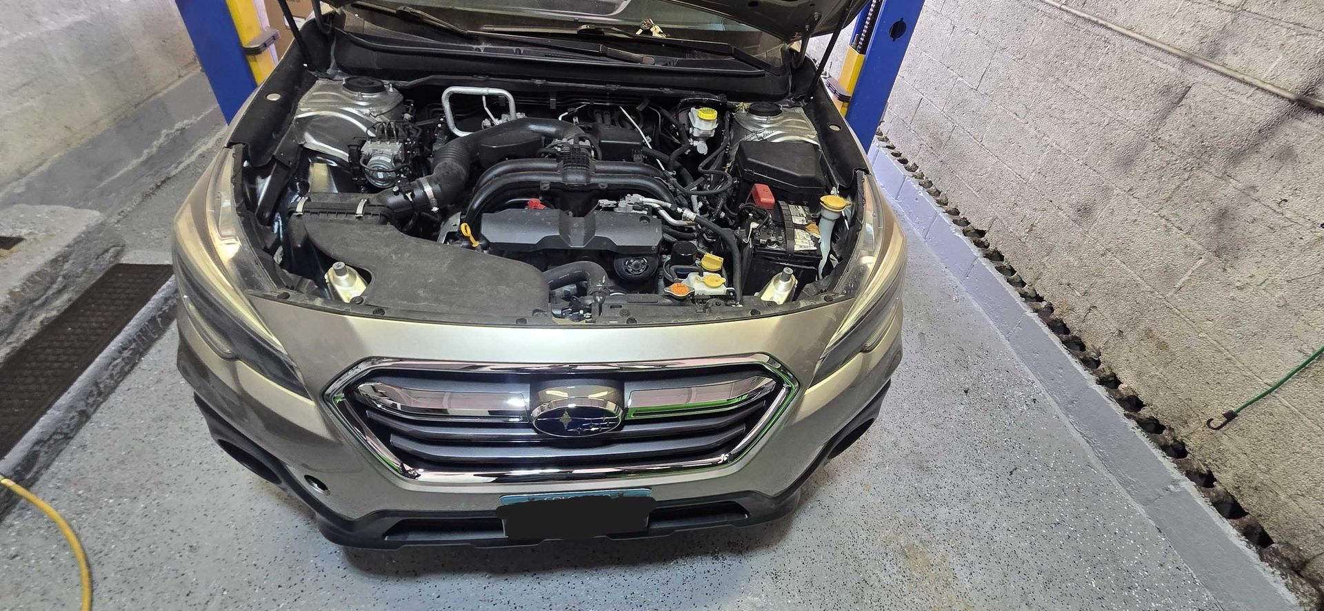 A silver Subaru Outback with its hood open, revealing the engine. The car is on a gray garage floor.