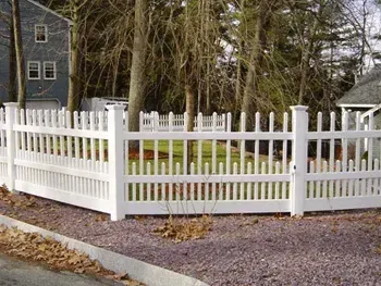 A white picket fence is surrounded by trees and gravel in front of a house.