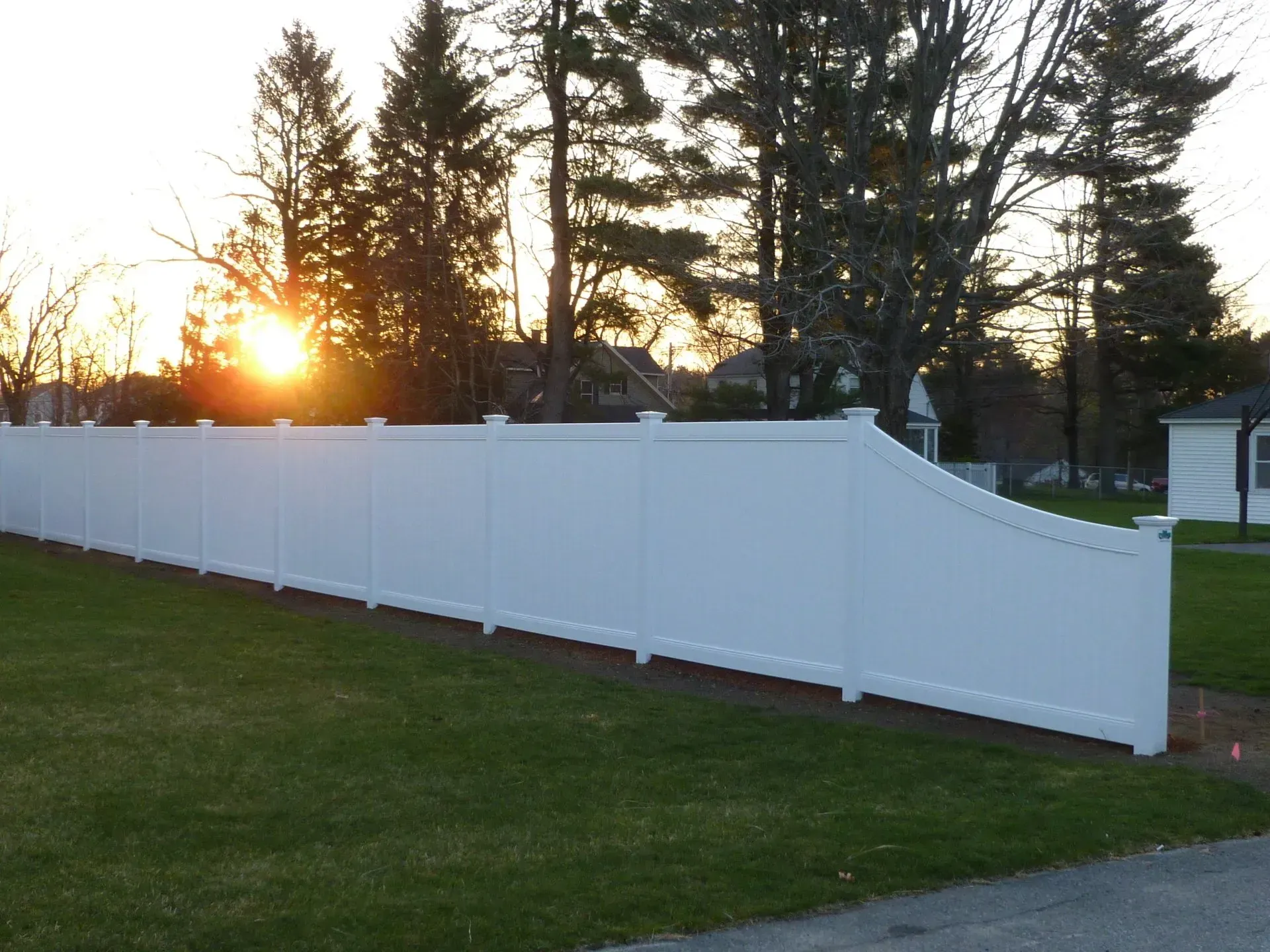 The sun is setting behind a white fence in a yard.