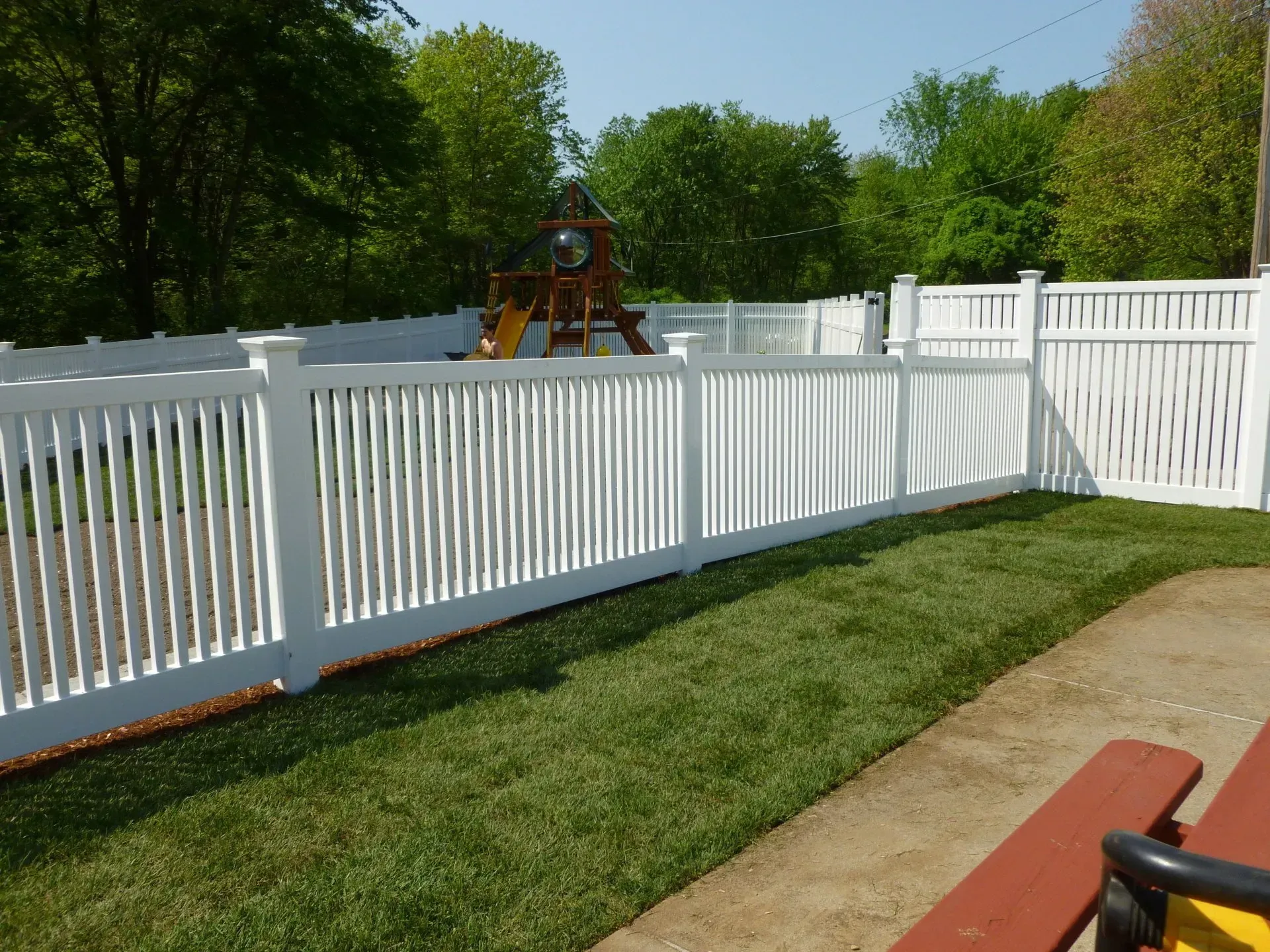 A white fence surrounds a backyard with a playground in the background