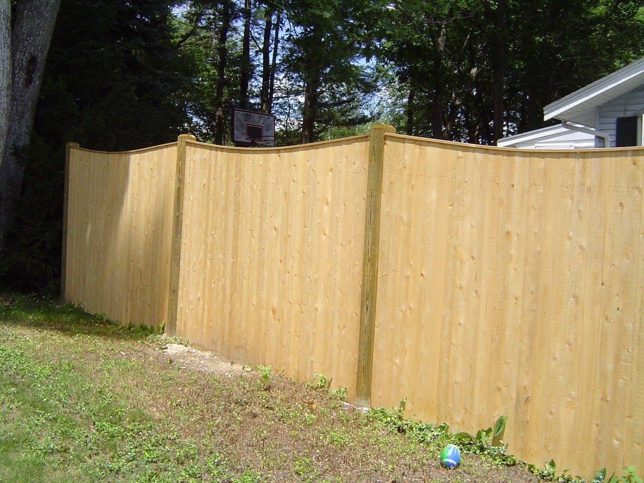 A wooden fence in a backyard with a basketball hoop in the background.