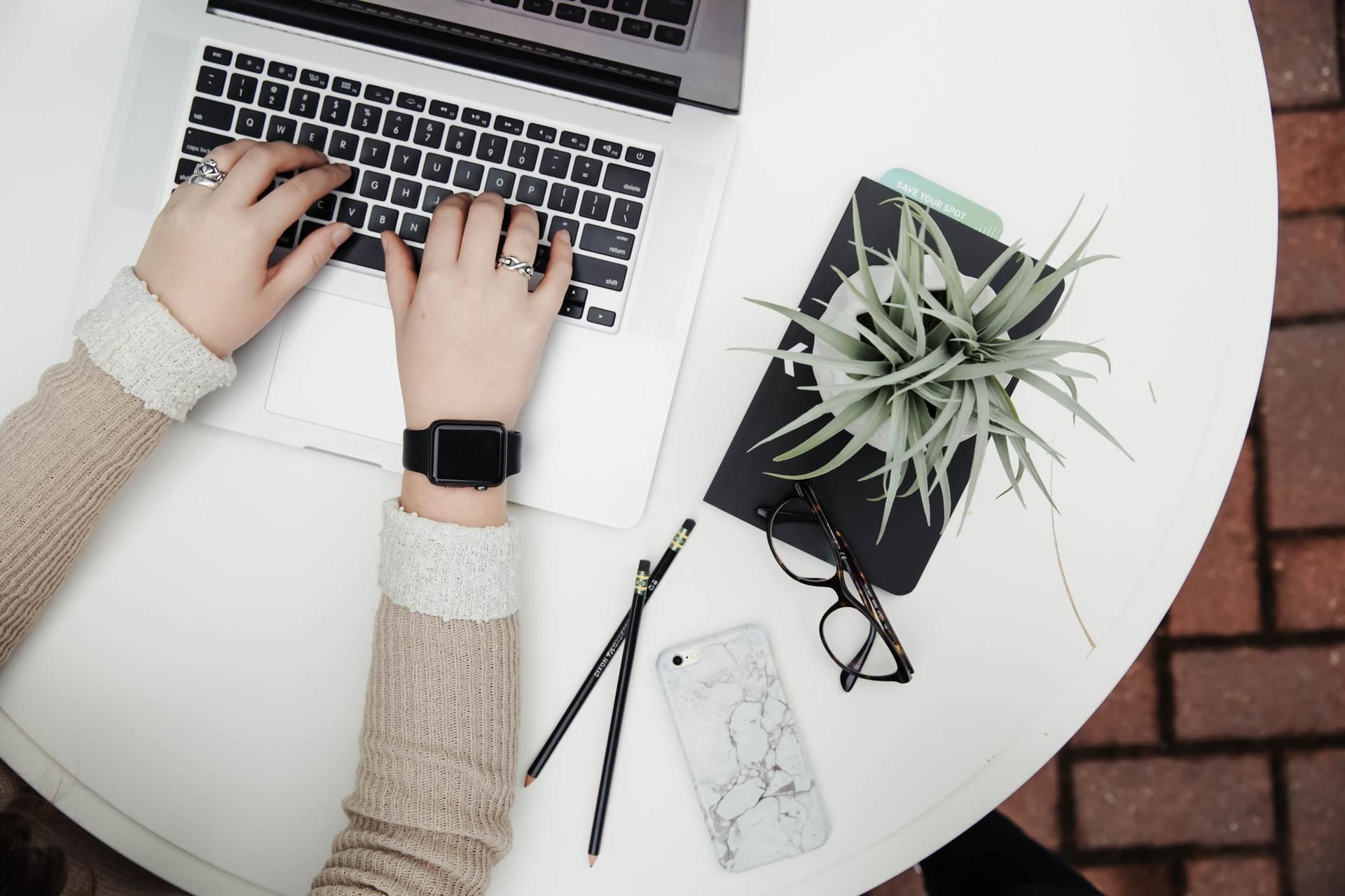 Hands typing on a laptop at a white table with a plant, notebook, glasses, pencil, and phone.