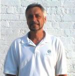 Man with graying hair and beard in a white polo shirt smiles in front of a white brick wall.