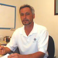 Man sitting at a desk, wearing a white polo shirt, in front of a whiteboard.