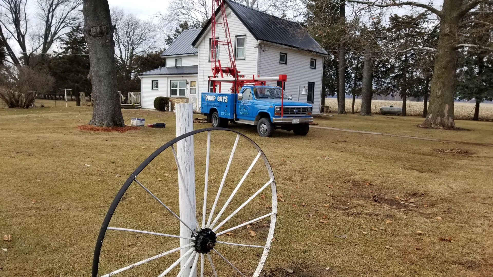 Blue truck with a crane in front of a white farmhouse on a grassy field, near a decorative wagon wheel.
