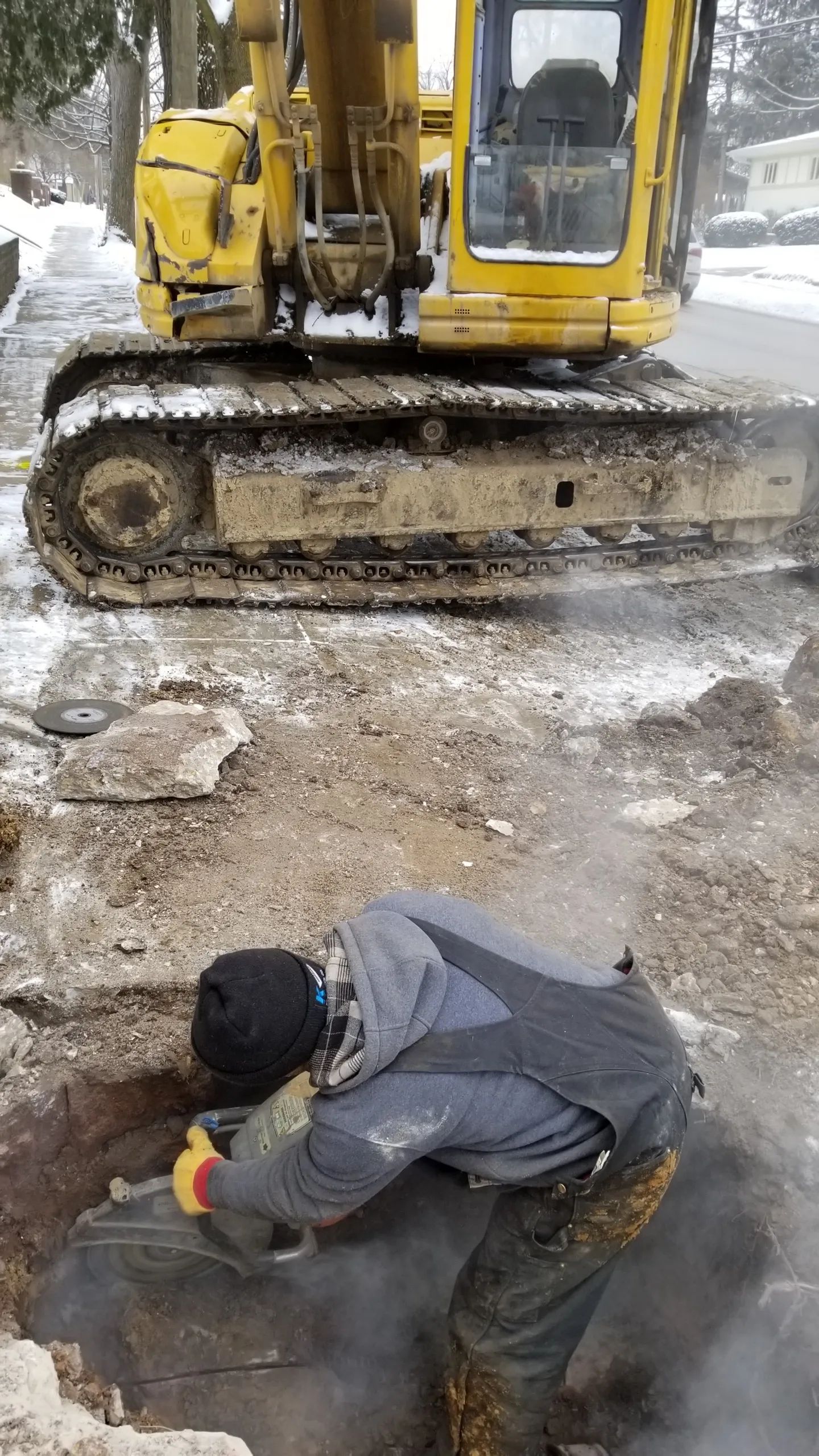 A person works in a trench near a yellow excavator in a snowy setting.