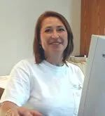 Woman smiling in a white t-shirt, seated near a computer monitor.