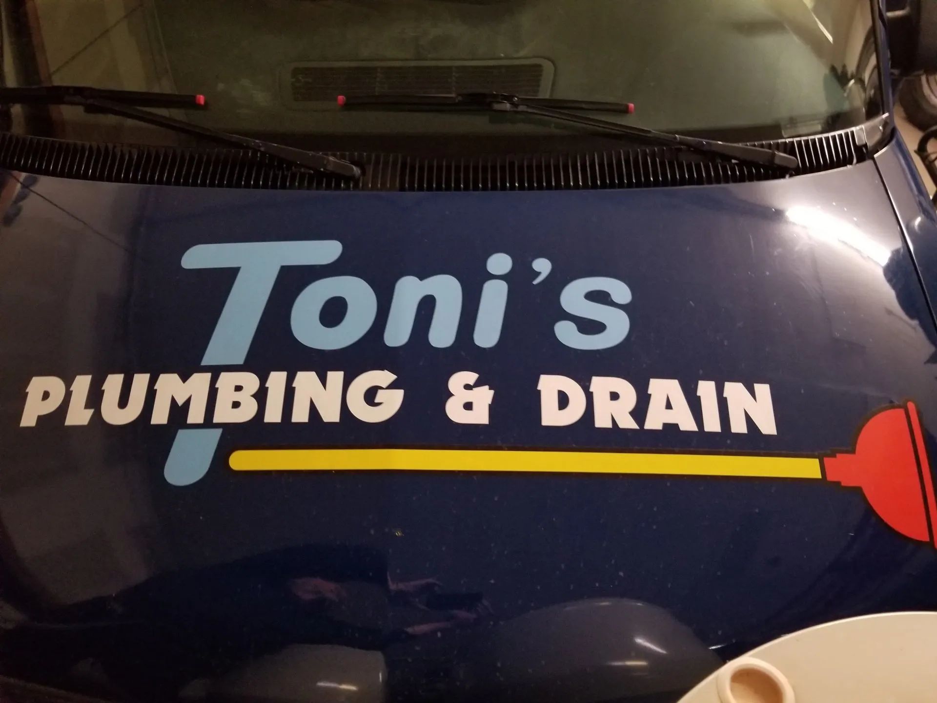 Toni's Plumbing & Drain logo on a blue van. Text is light blue and white. Graphic of a plunger, yellow handle, red head.