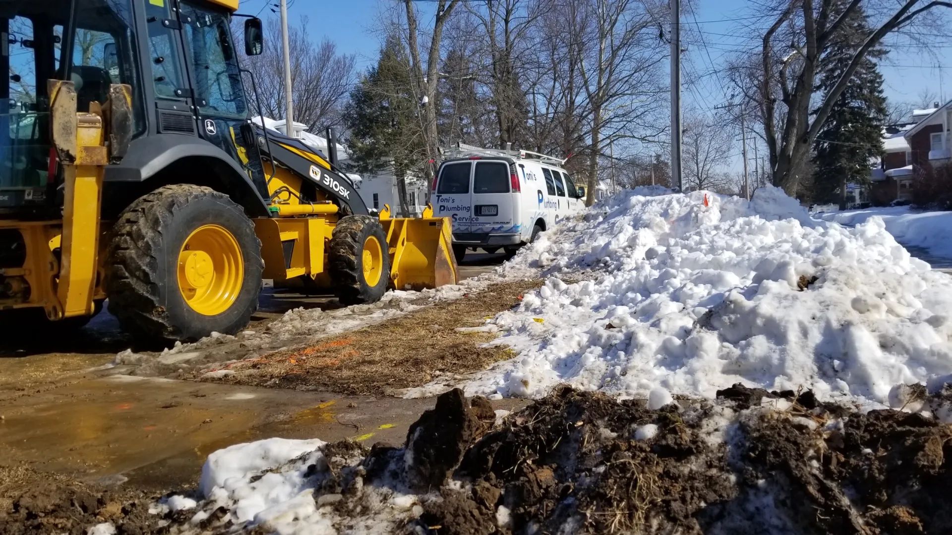 Yellow backhoe clearing snow from a street, next to a white van on a sunny day.