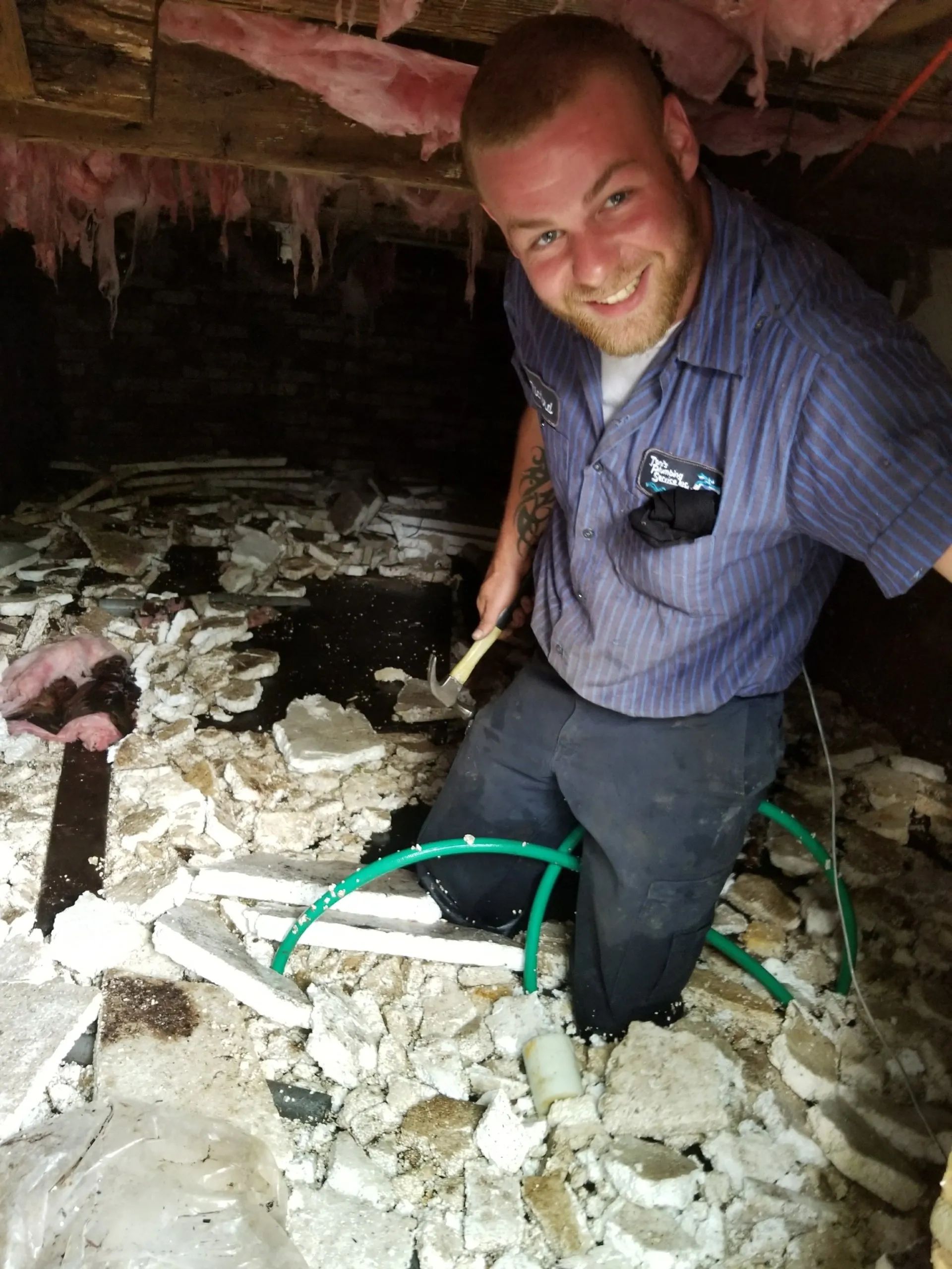 A person in blue work shirt smiles while working in a crawlspace with debris and green tubing.