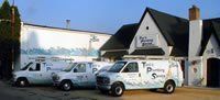 Three white plumbing service vans parked outside a white building with black roof and chimney.