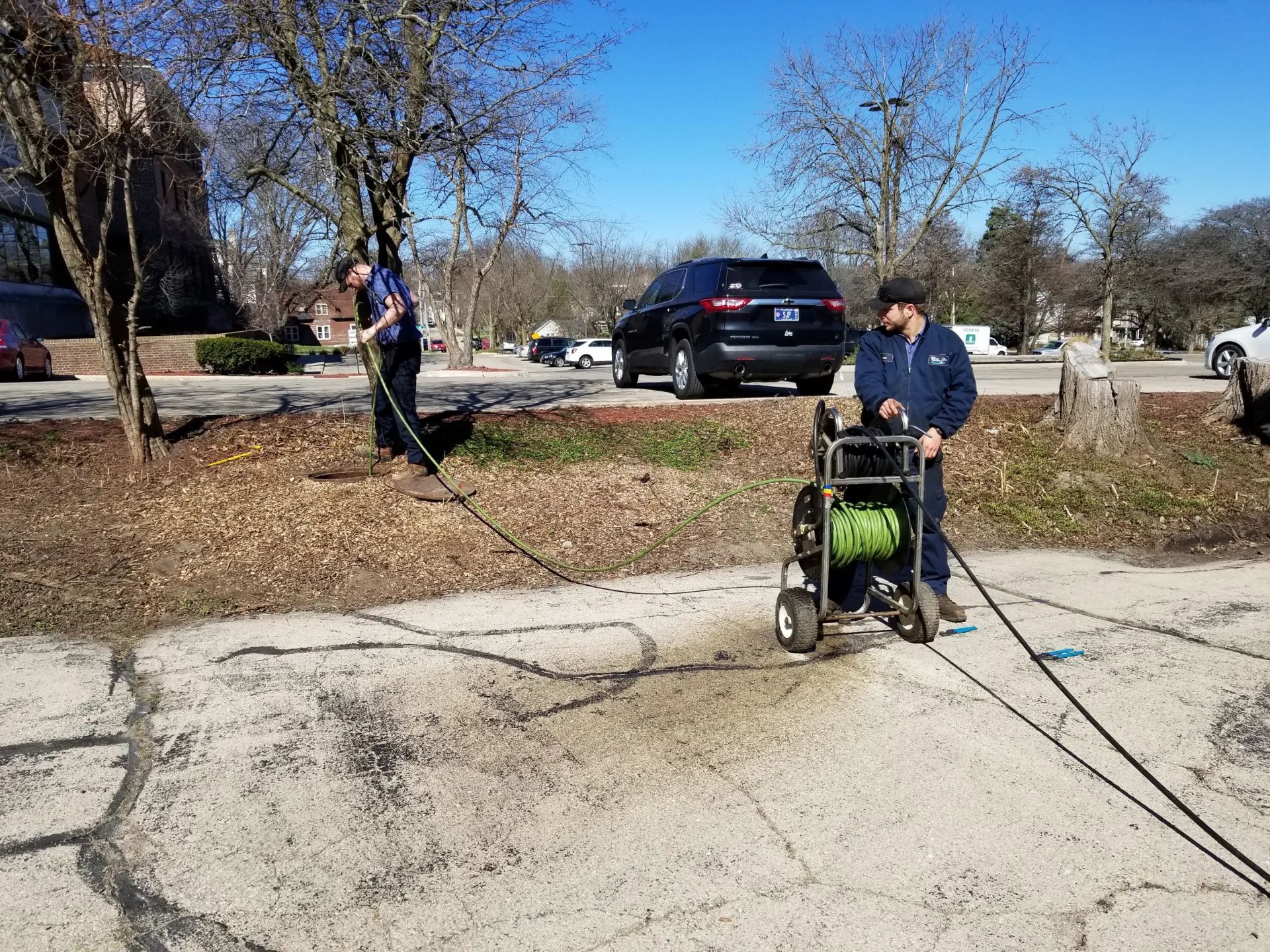 Two people pressure washing a cracked asphalt lot, near trees and parked vehicles.
