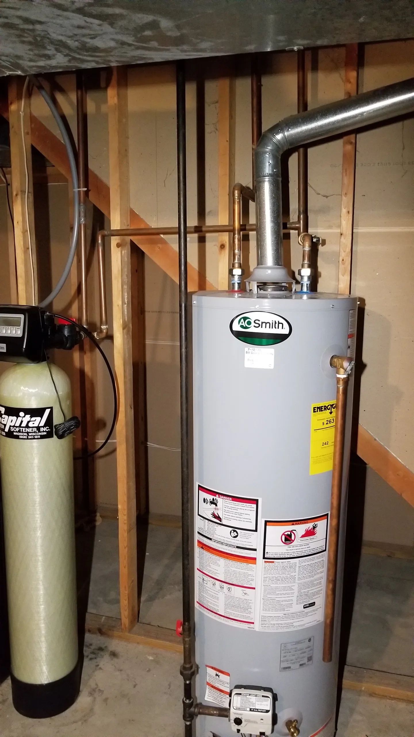 Water heater and water softener in a basement setting. The heater is cylindrical and metal; the softener is tall and beige.