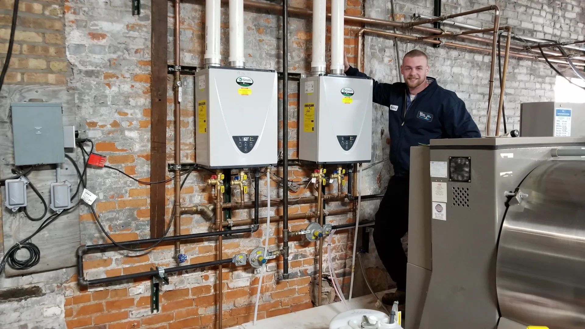 Plumber smiles next to two wall-mounted water heaters with copper pipes, set against a brick wall.