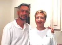 Man and woman smiling, indoors. Both wear white shirts, standing close, arms touching.
