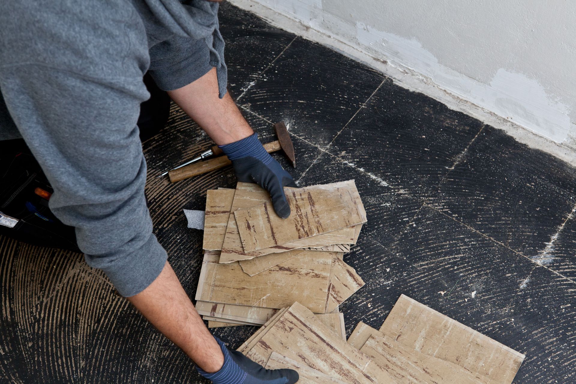 Person in gray hoodie removing floor tiles with hammer.