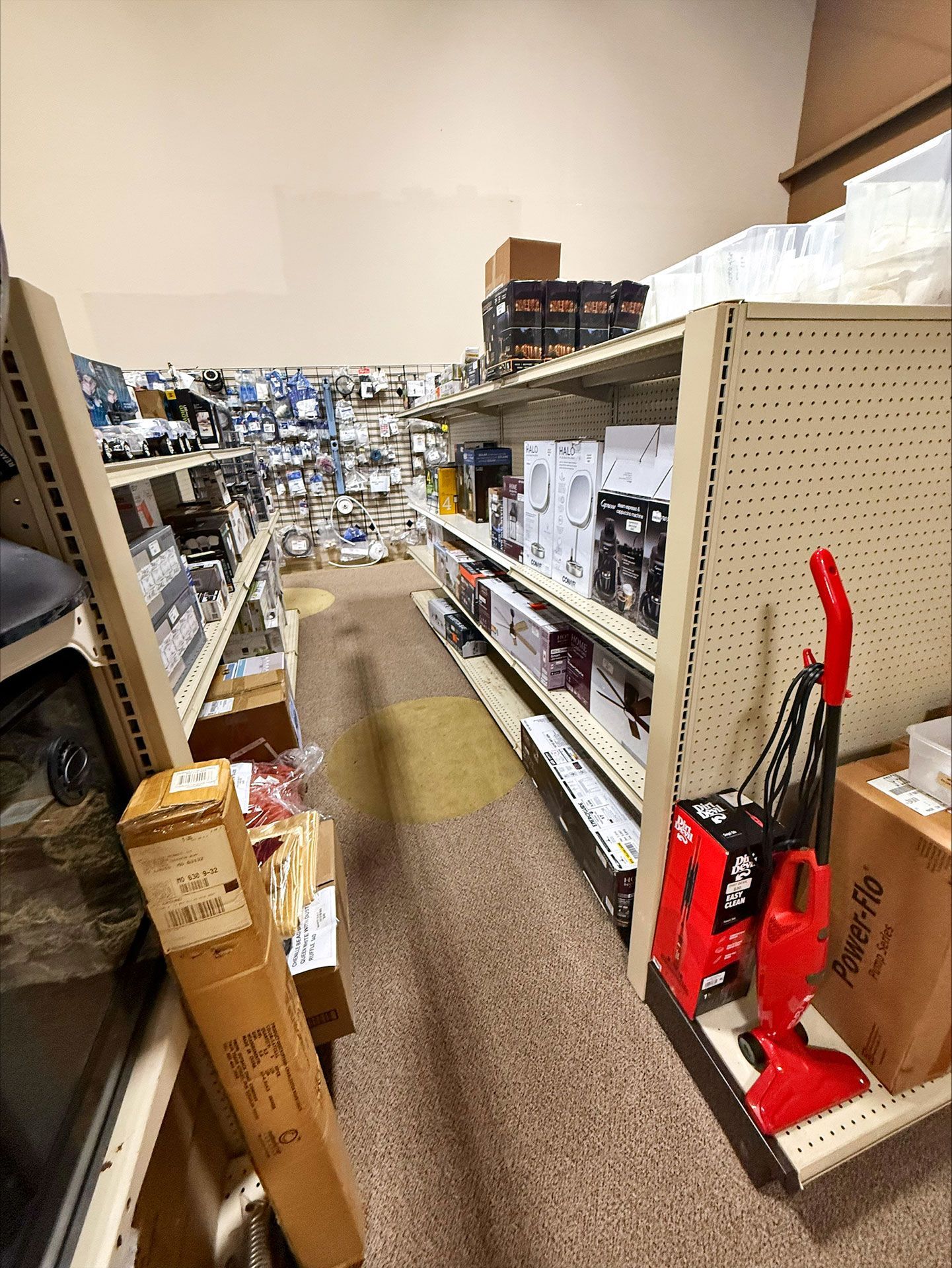 Narrow store aisle lined with shelves displaying various products, a vacuum cleaner is prominent.