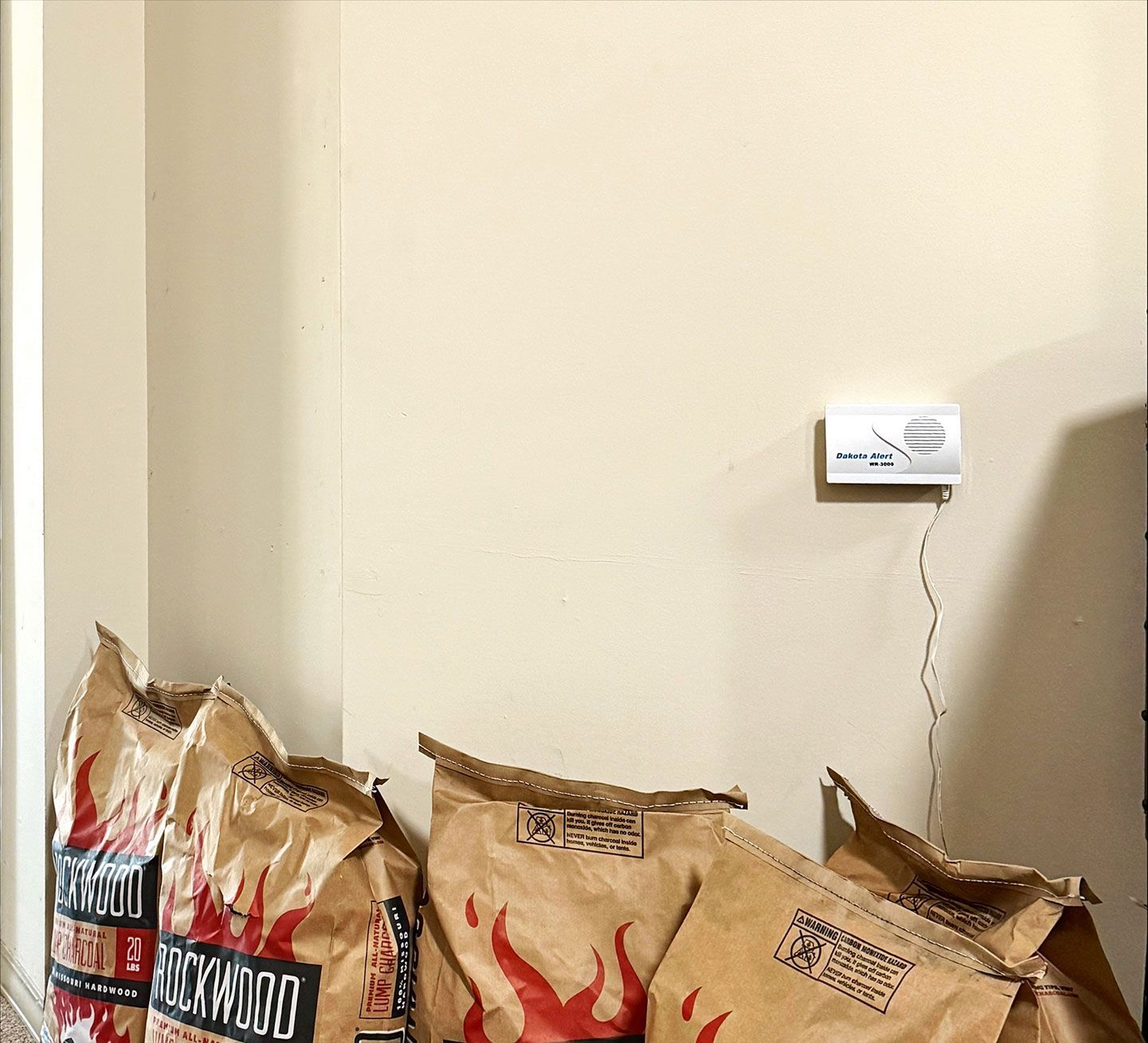 Bags of charcoal stacked against a beige wall with a smoke detector.