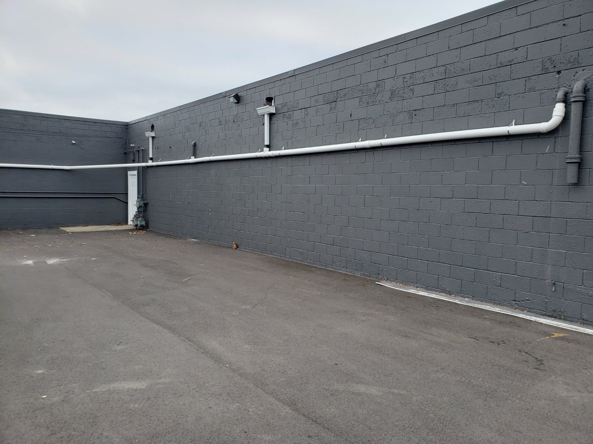 Gray brick wall with white pipes running horizontally, and asphalt ground.