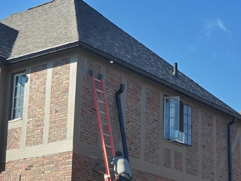 Person on a ladder installing a black downspout on a brick building with a dark roof.