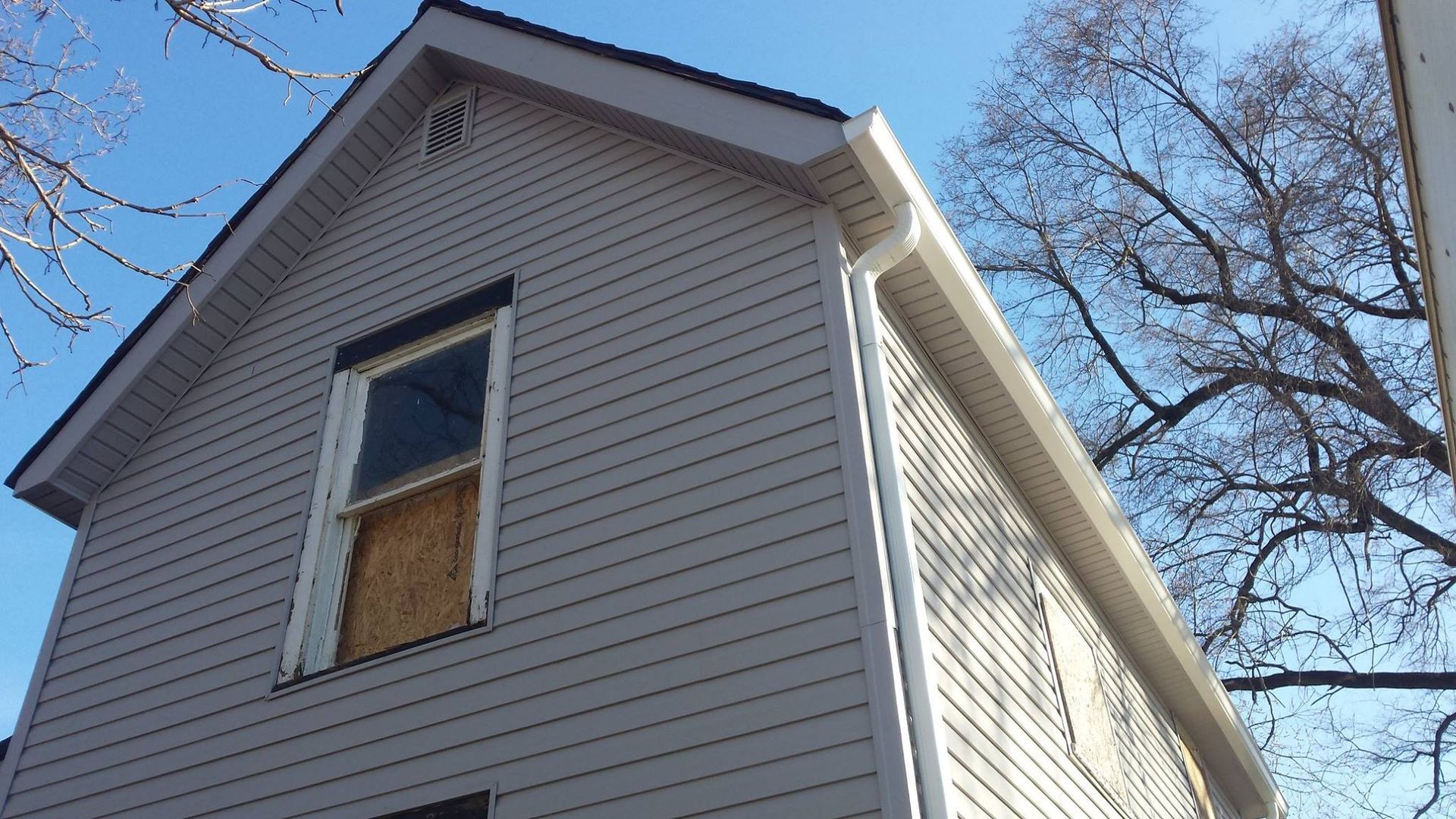 Two-story house with gray siding, boarded-up window, and white gutters against a clear blue sky.