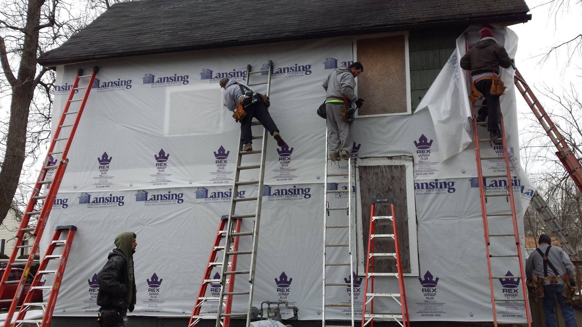 Workers install house siding using ladders. The house is partially covered in white wrap.