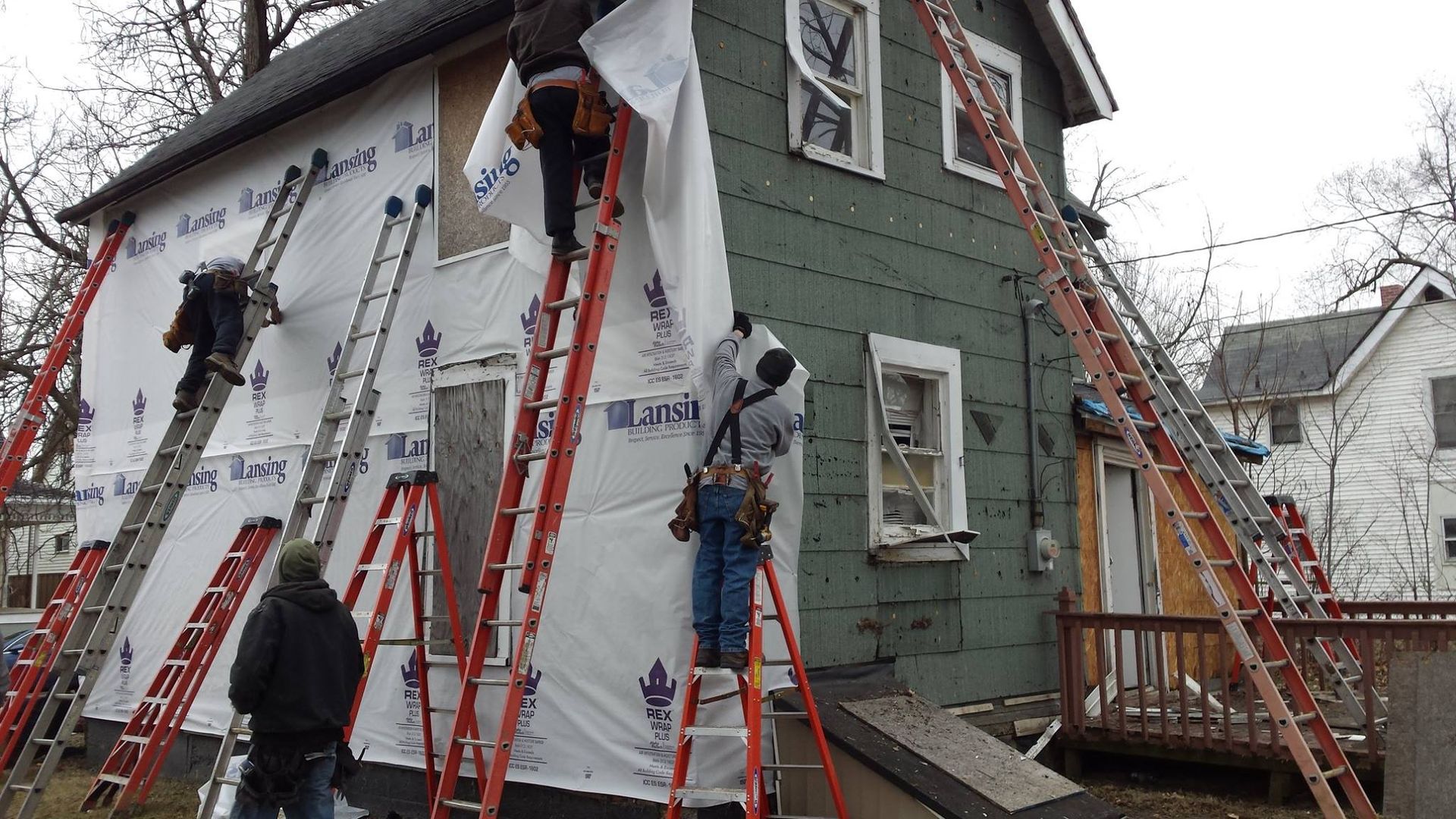 Workers on ladders installing white sheathing on the exterior of a two-story green house.