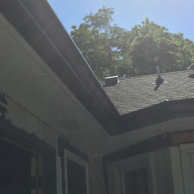 Gutters on a house with a dark roof and trees in the background under a blue sky.