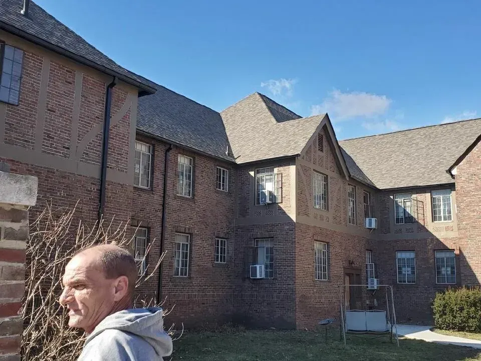 Brick apartment building with a man on the left. Blue sky, grass, and bare bush in front.