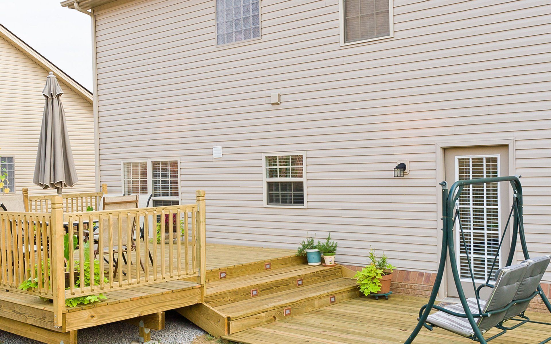 Wooden deck and steps leading to a two-story beige house with a swing, umbrella, and furniture.