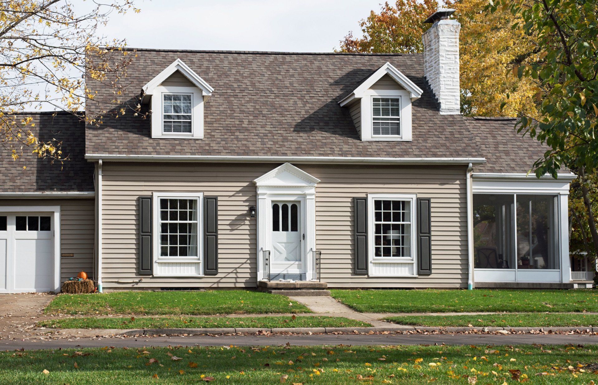 Tan house with white trim, two dormer windows, shutters, chimney, and screened porch on a green lawn.
