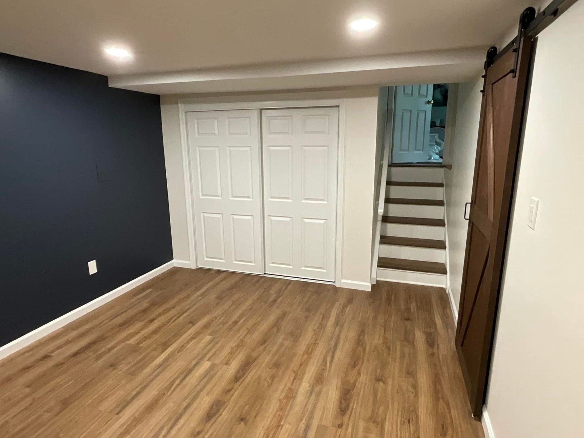 Empty room with wood floor, dark accent wall, white closet doors, and stairs beside a doorway.