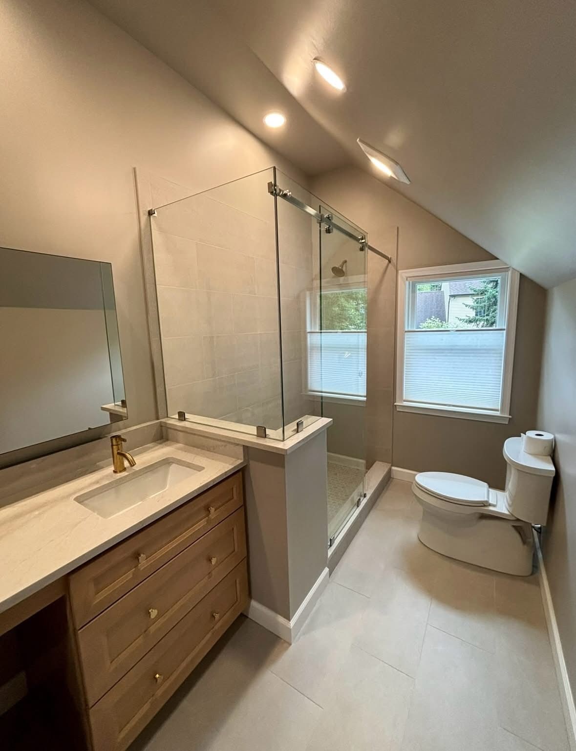 Bright attic bathroom with white tub, wood vanity, toilet, and two windows under sloped ceiling.
