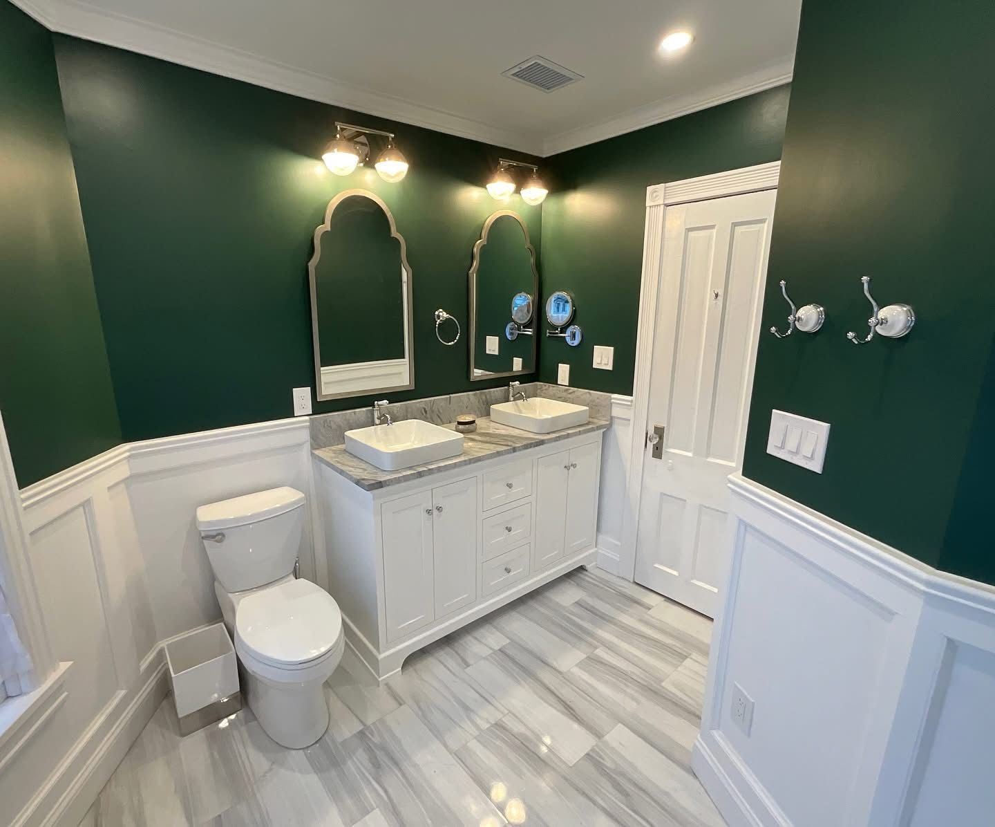 Modern bathroom with dark green walls, white vanity, toilet, double mirrors, and marble-look floor.