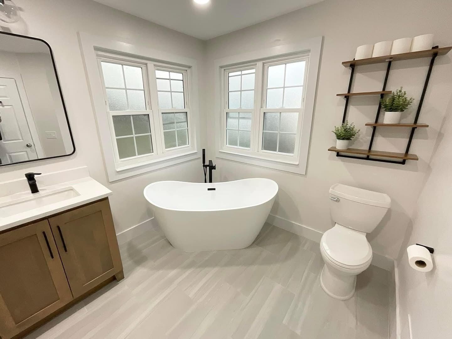 Bright modern bathroom with freestanding tub, vanity, toilet, windows, and wall shelves
