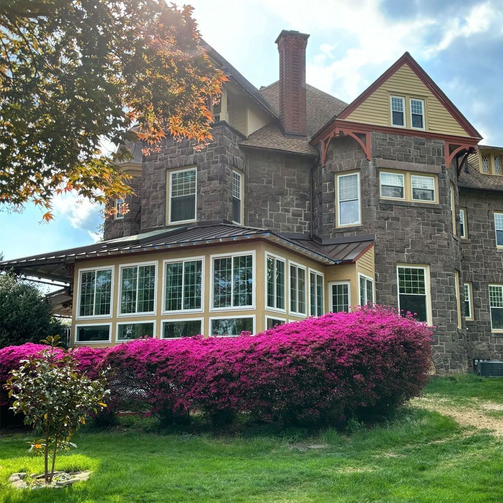 Stone house with large windows and pink flowering hedge in a sunny green garden