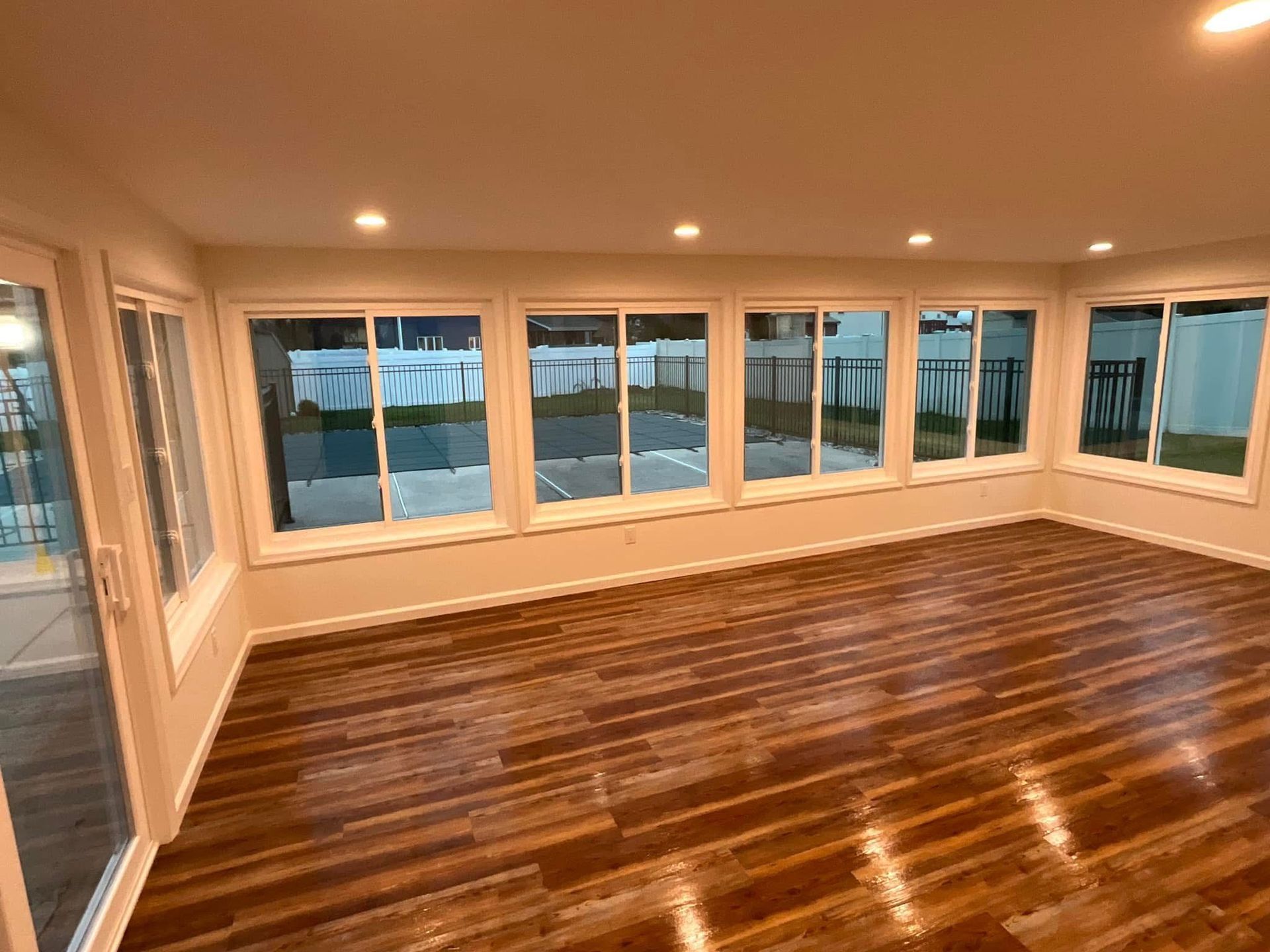Empty sunroom with hardwood floors, recessed lights, and large windows overlooking water.