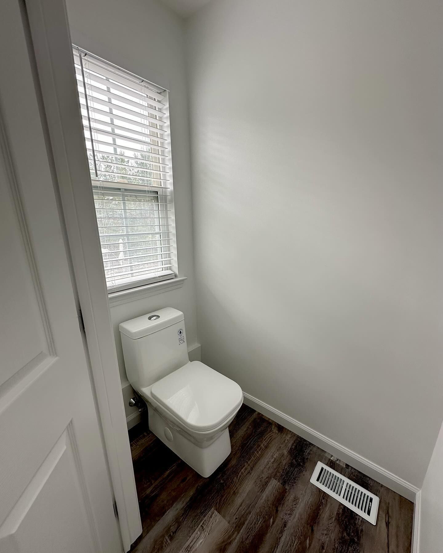 Small bathroom with a white toilet, window with blinds, and a floor vent on dark wood flooring.