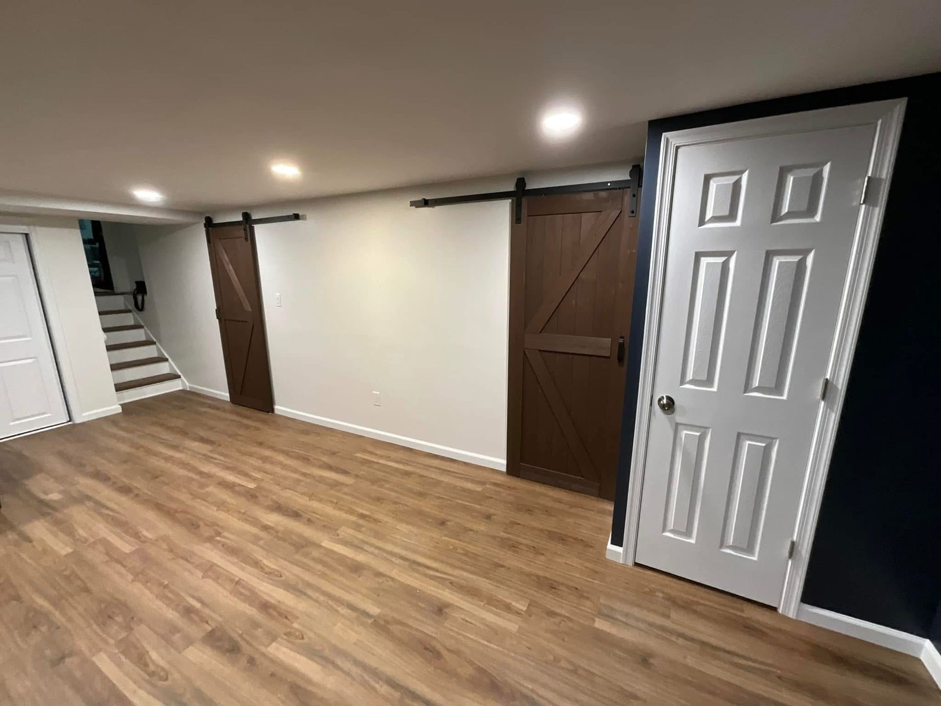 Empty finished basement with wood floor, white walls, brown barn doors, and a stairway on the left.