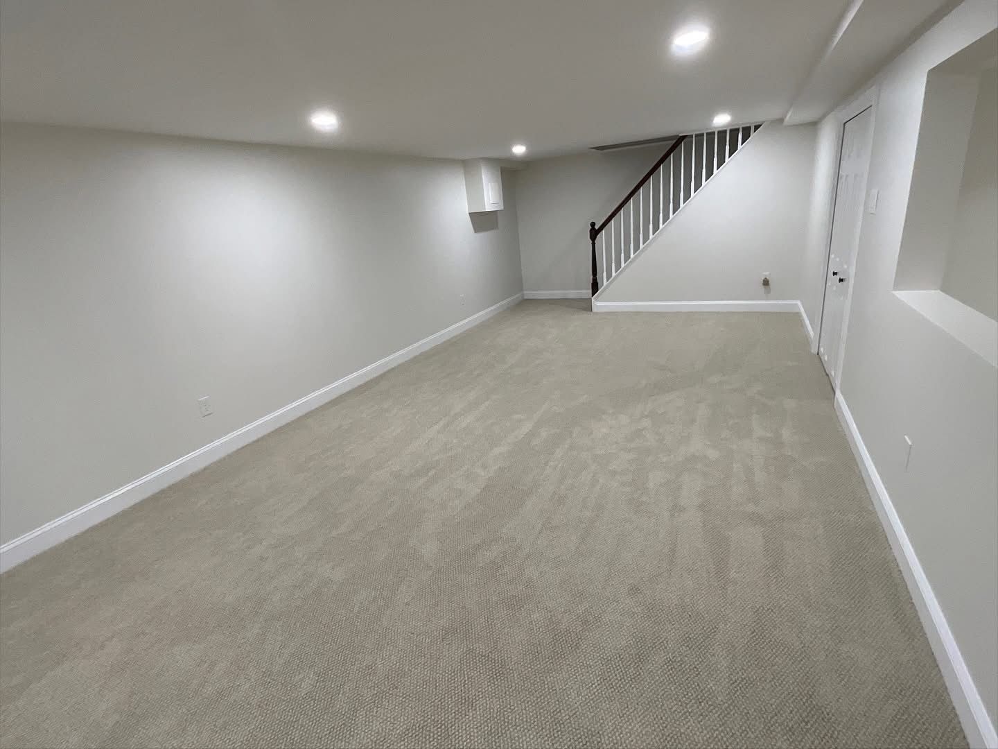 Empty carpeted basement room with white walls, recessed lights, and staircase at the far end.