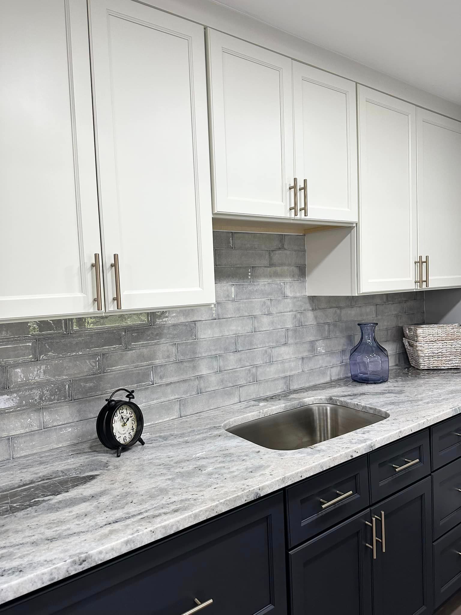 Modern kitchen with white cabinets, gray stone backsplash, dark lower cabinets, and stainless steel sink.