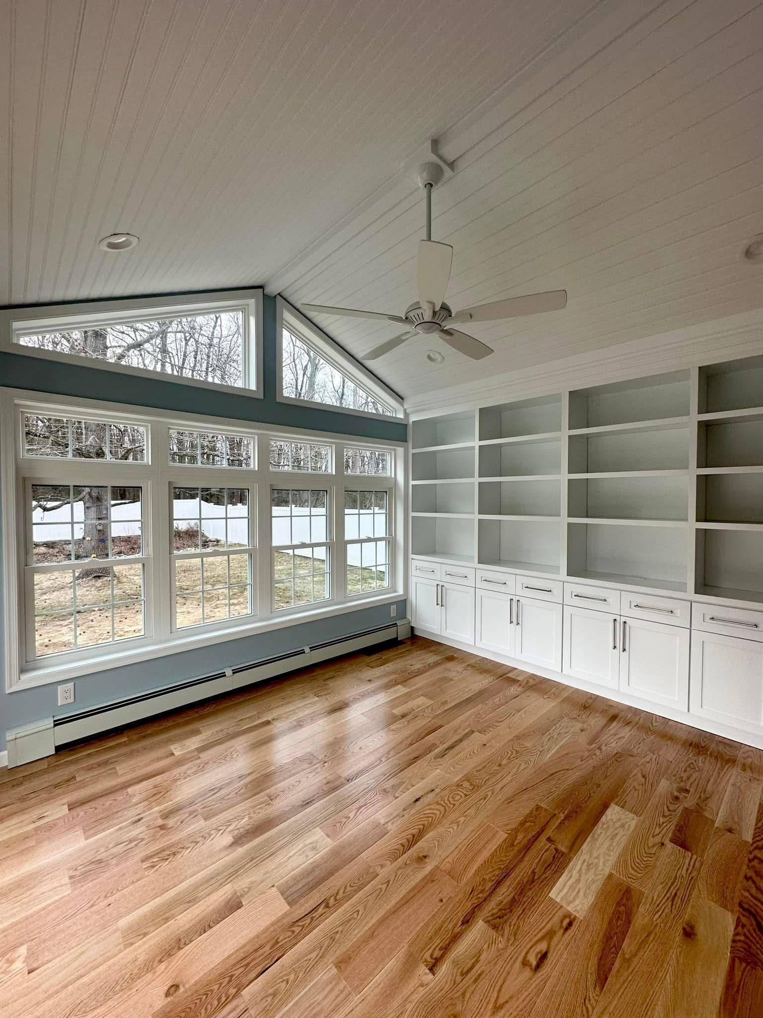 Bright empty sunroom with hardwood floors, white built-in shelves, and large windows under a vaulted ceiling.