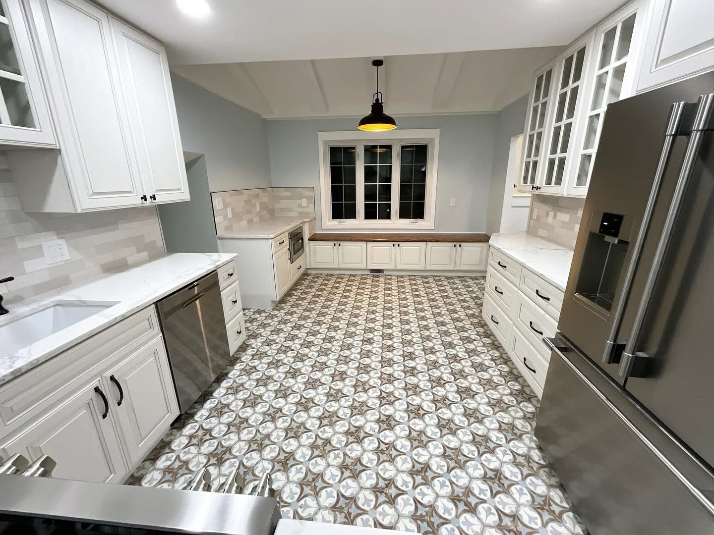Bright white kitchen with patterned tile floor, stainless fridge, and window seat beneath a window.