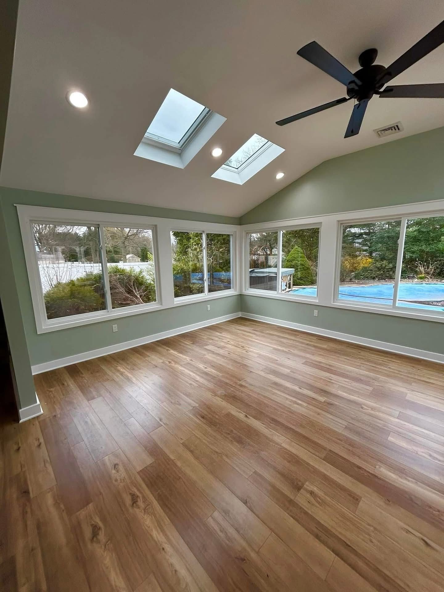 Bright sunroom with hardwood floors, skylights, ceiling fan, and large windows overlooking a pool and greenery.
