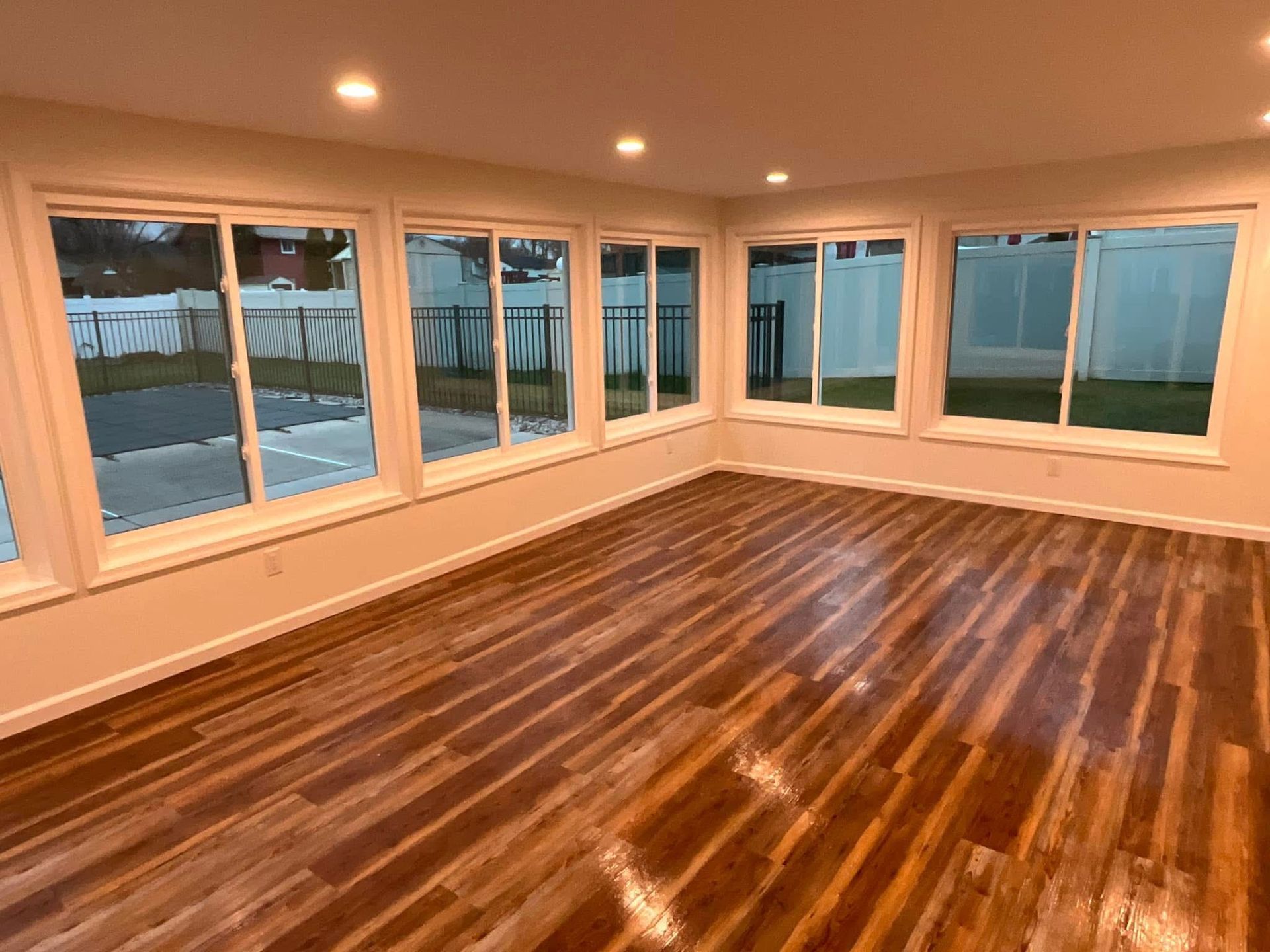 Empty sunroom with glossy hardwood floors, white walls, and large windows overlooking a snowy yard.